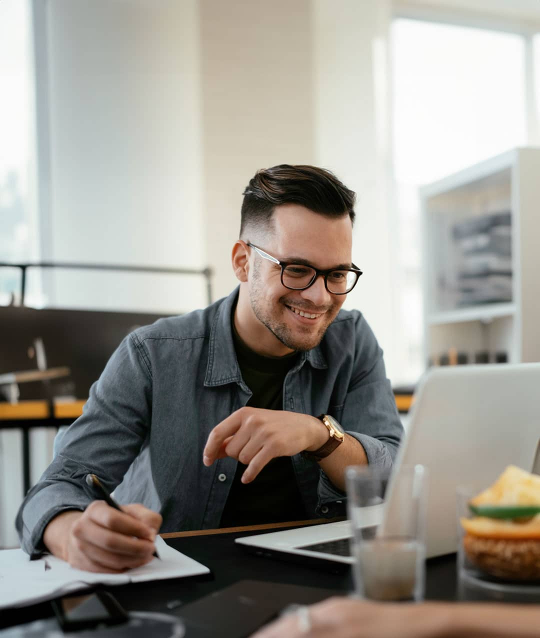 Lächelnder Mann mit Brille arbeitet an einem Laptop und notiert Informationen, repräsentiert die Planung und Umsetzung sicherer digitaler Prozesse für eine 27001-Zertifizierung.